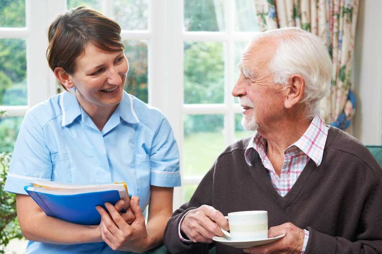 An older couple smiles and holds hands in a cozy, sunlit apartment living room with personal decorations.