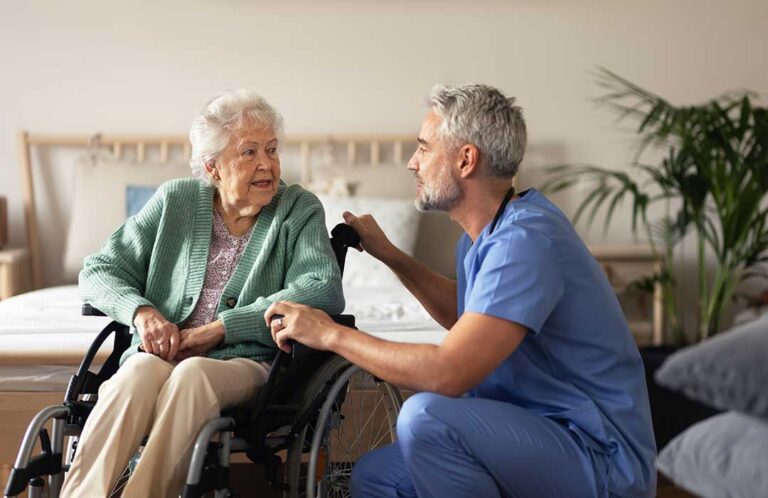 Caregiver assists an older resident in a bright assisted living hallway while another staff member reviews a care plan.